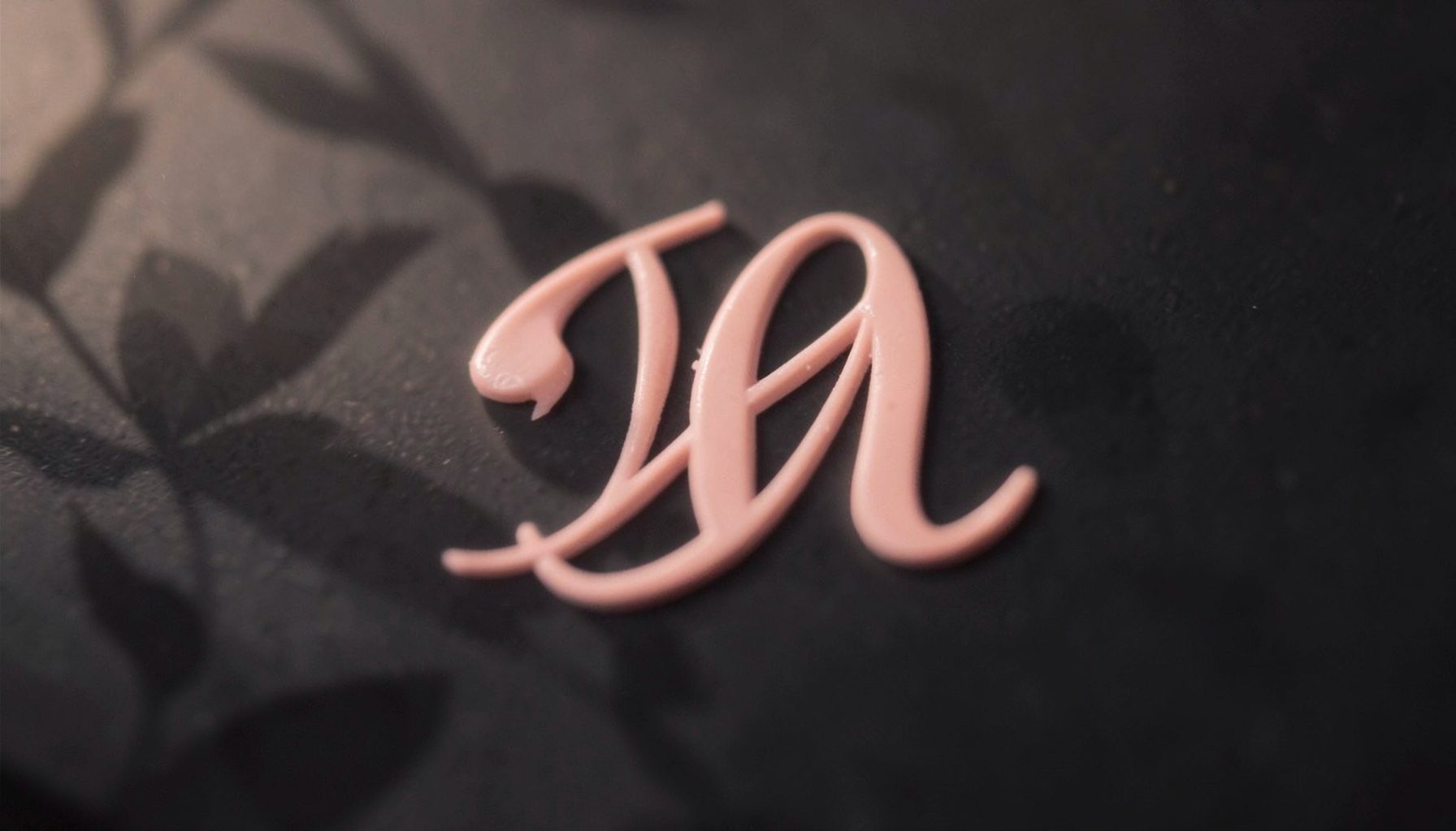 A soft, dreamy close-up of a blush-pink monogram letter on a dark charcoal background, with delicate floral shadows.