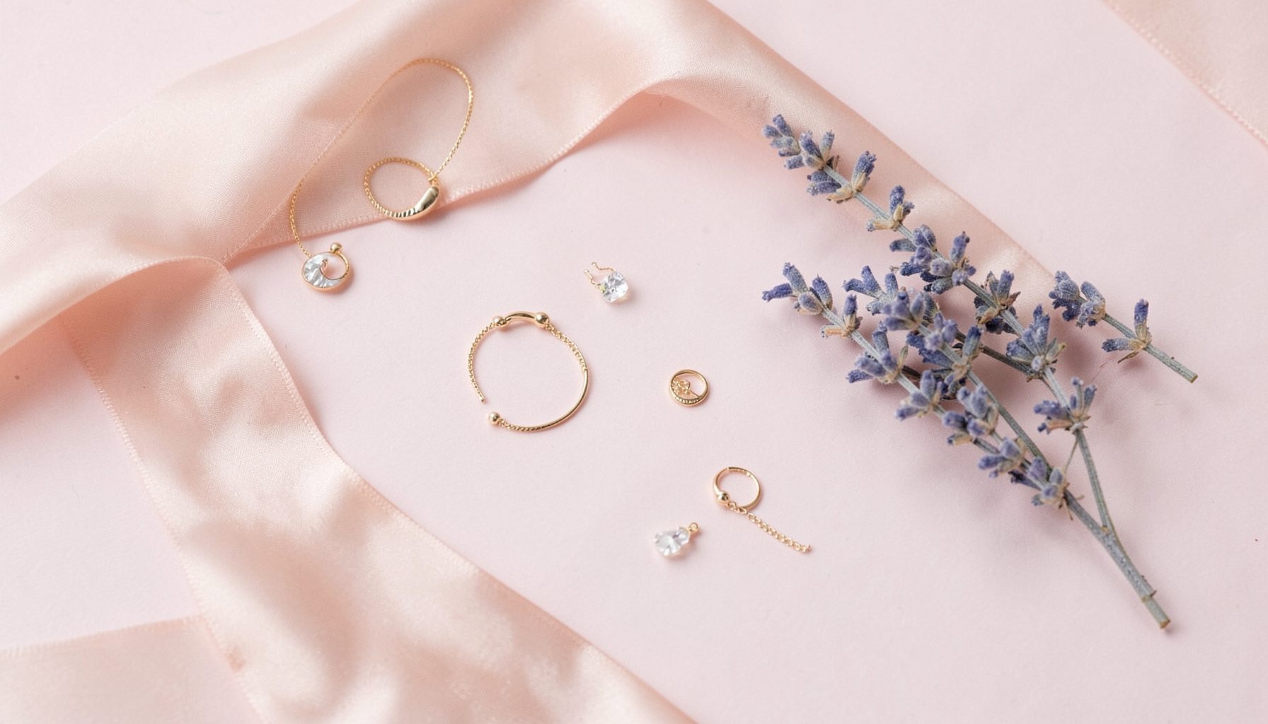 Softly lit flat-lay on a pale pink surface with delicate gold jewelry, a blush-toned silk ribbon, and scattered feminine accents