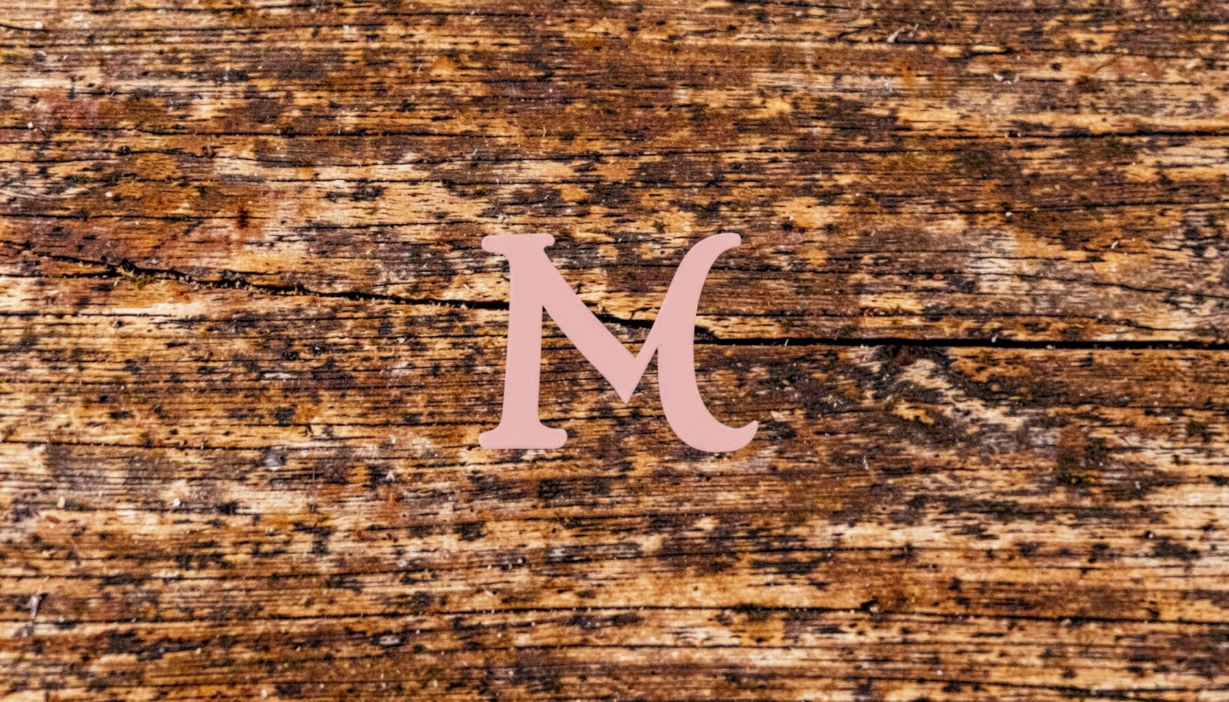 Warm rustic wood textures in deep browns and soft creams, with a subtle monogram letterform in muted blush pink, evoking a cozy design-studio feel.
